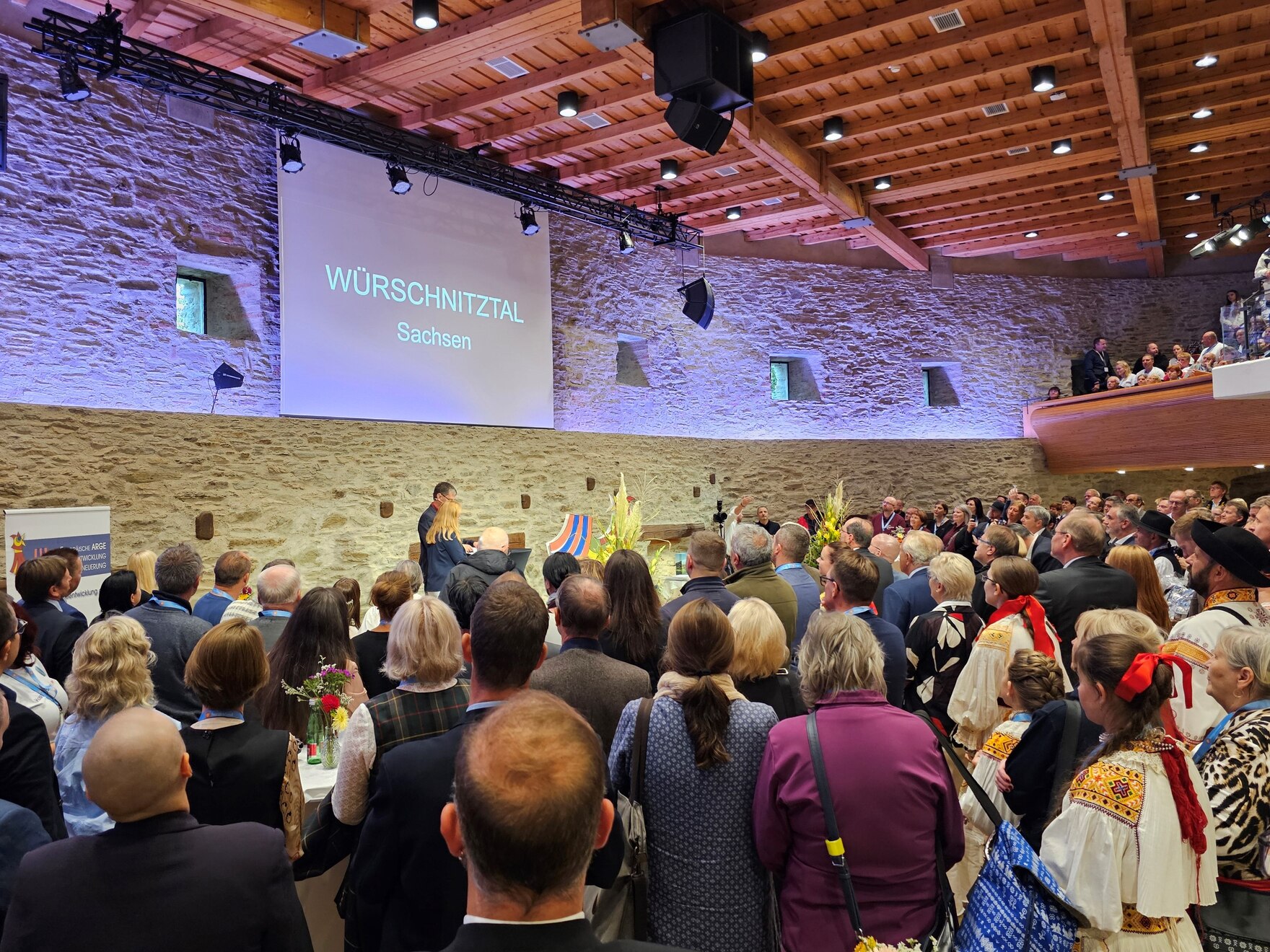 Eine Menge Leute in einem Saal – im Hintergrund eine große Leinwand mit der Projektion Würschnitztal Sachsen