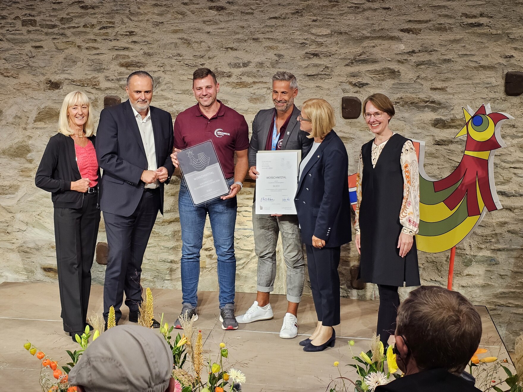 Sechs Personen stehen auf einer Bühne, in der Mitte die zwei Bürgermeister mit Plakette und Urkunde