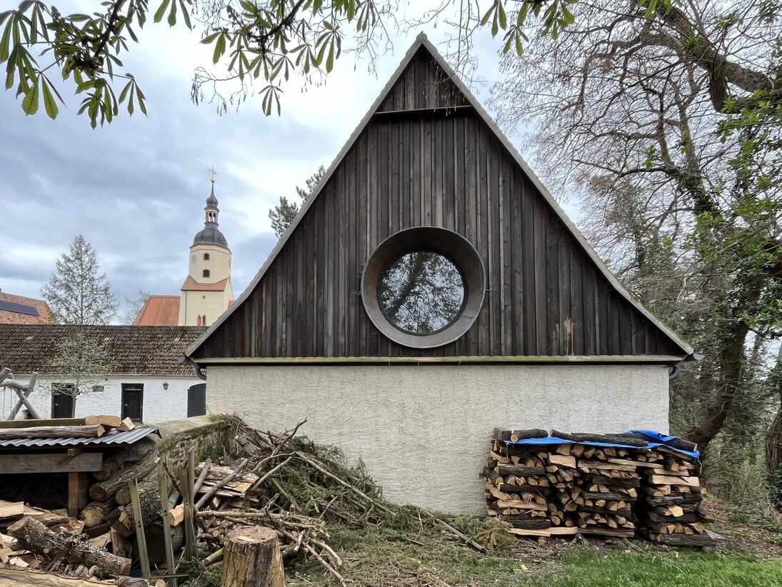 Giebelansicht mit Rundfenster, davor Holzstapel, dahinter der Kirchturm