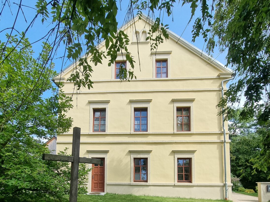 Sicht auf die beige Giebelseite mit Holzkreuz im Vordergrund