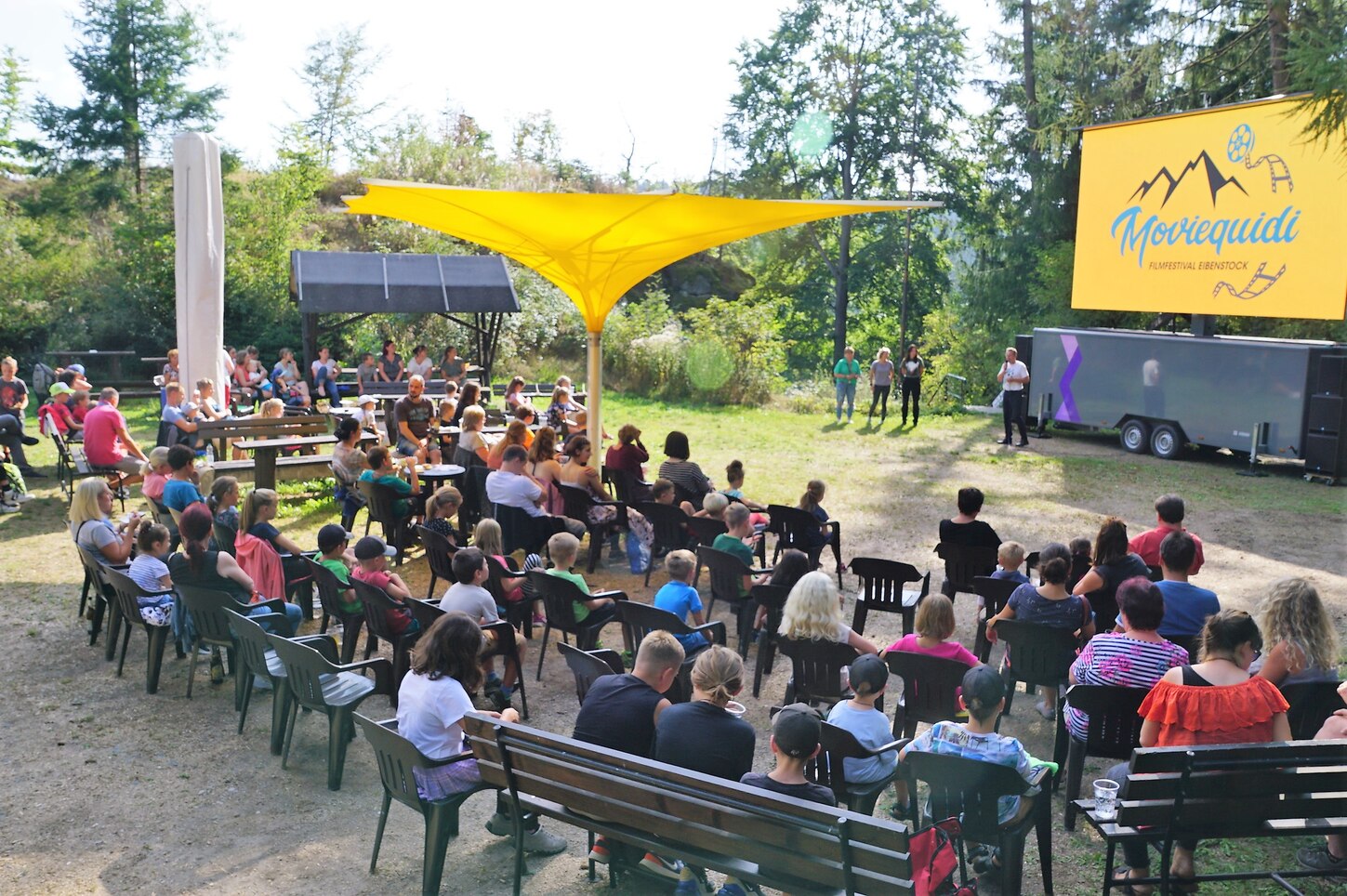 im Vordergrund mehrere Sitzreihen mit Gästen auf dem Platz im Freien; im Hintergrund eine aufgebaute Leinwand mit der Aufschrift Moviequidi