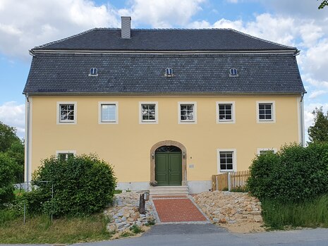 Zweigeschossiger Bau mit Doppelwalmdach mit Sträuchern vorm Hauseingang